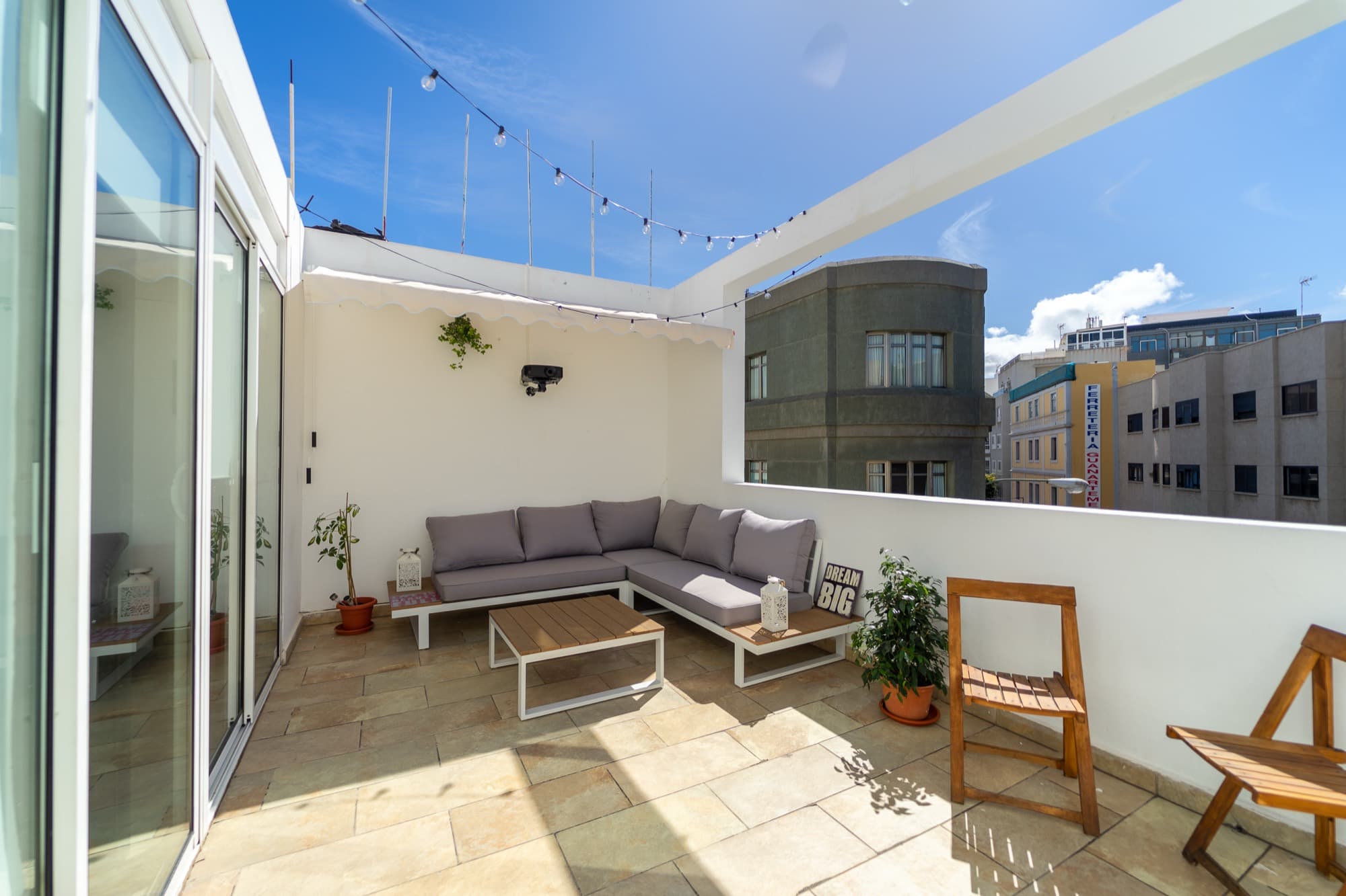 La terraza de House Canteras al sol: sofá exterior gris, sillas plegables de madera y guirnaldas de luces bajo el cielo azul.